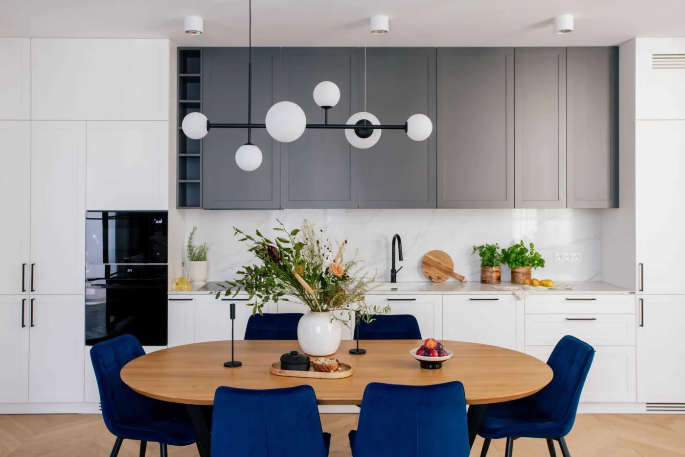 Dining area with an oval table and blue upholstered chairs beside a white and grey kitchen